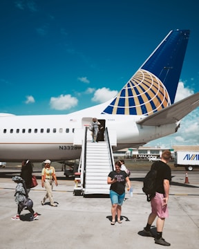 A large commercial airplane is parked on a tarmac under a clear blue sky. People are descending a mobile staircase from the aircraft, carrying bags and wearing casual attire. The airplane has a distinctive blue and gold tail fin design. The background includes airport buildings and a service vehicle.