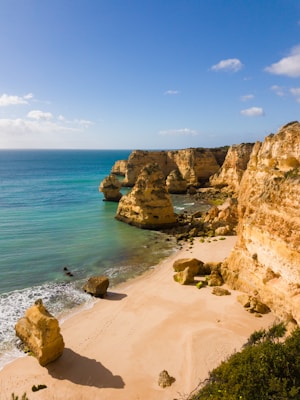 A serene beach scene with golden sandy shores and towering limestone cliffs. The azure waves gently lap against the shore under a bright blue sky dotted with a few fluffy clouds.