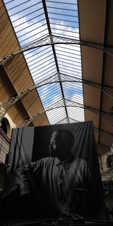 A black and white portrait of a man looking contemplatively through a window, displayed in a large building with a glass ceiling. The structure of the building includes metal beams and arches, creating a spacious and airy atmosphere. The sky can be seen through the glass roof, adding a sense of openness.