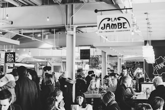 A bustling indoor market filled with people, featuring various food and other stalls. Signs for options like 'La Jambe' offering wine, cheese, and charcuterie, and 'Zita Rica' are visible. The atmosphere is lively and social, with groups chatting and dining.