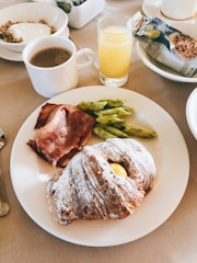 A breakfast setting with a plate featuring a dusted pastry, slices of bacon, and a small serving of seasoned penne pasta. To the side, there is a cup of coffee and a glass of orange juice. In the background, a bowl of granola with yogurt and a packaged granola bar add to the meal.