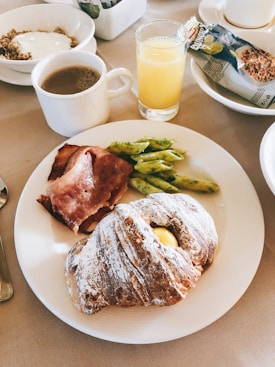 A breakfast setting with a plate featuring a dusted pastry, slices of bacon, and a small serving of seasoned penne pasta. To the side, there is a cup of coffee and a glass of orange juice. In the background, a bowl of granola with yogurt and a packaged granola bar add to the meal.