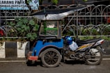 A blue motorized tricycle with a canopy parked on a street. The vehicle is adjacent to a curbside garden with green plants and is positioned in front of a metal fence. A blue helmet rests on the motorcycle seat.
