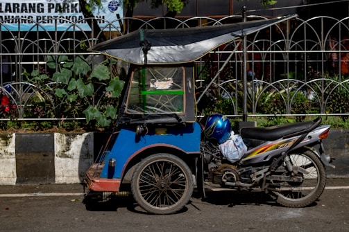 A blue motorized tricycle with a canopy parked on a street. The vehicle is adjacent to a curbside garden with green plants and is positioned in front of a metal fence. A blue helmet rests on the motorcycle seat.