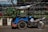 A blue motorized tricycle with a canopy parked on a street. The vehicle is adjacent to a curbside garden with green plants and is positioned in front of a metal fence. A blue helmet rests on the motorcycle seat.