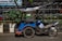 A blue motorized tricycle with a canopy parked on a street. The vehicle is adjacent to a curbside garden with green plants and is positioned in front of a metal fence. A blue helmet rests on the motorcycle seat.