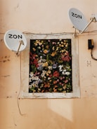 A wall with two satellite dishes mounted on either side of a window adorned with a vibrant array of artificial flowers in various colors and types. The surface of the wall is a worn, pale peach color, and an electrical box is affixed to the wall near the right dish.