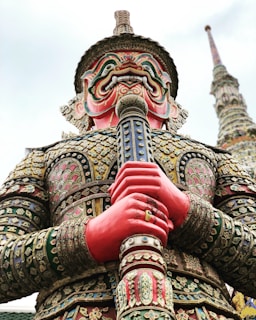 A large, ornate statue of a mythological figure with a fierce, colorful face and intricate armor. The statue features elaborate patterns and details in its clothing, and it is holding a large staff or weapon. The background shows a tall, decorated spire extending into the sky.