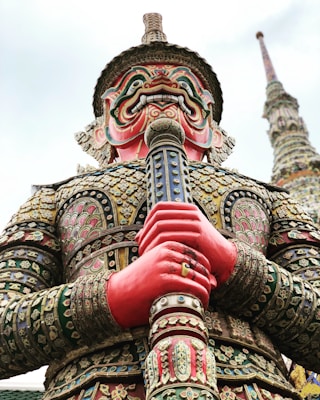 A large, ornate statue of a mythological figure with a fierce, colorful face and intricate armor. The statue features elaborate patterns and details in its clothing, and it is holding a large staff or weapon. The background shows a tall, decorated spire extending into the sky.