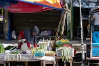A person wearing a headscarf is tending to a variety of vegetables displayed on a wooden cart in a street market. The produce includes eggplants, green onions, and long beans, all organized in white plastic crates. The market stall is shaded by a makeshift orange and blue tarpaulin. A closed shop with a shutter is in the background.