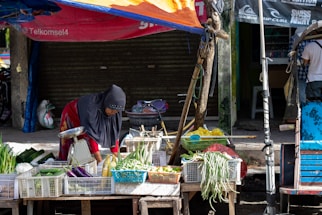 A person wearing a headscarf is tending to a variety of vegetables displayed on a wooden cart in a street market. The produce includes eggplants, green onions, and long beans, all organized in white plastic crates. The market stall is shaded by a makeshift orange and blue tarpaulin. A closed shop with a shutter is in the background.