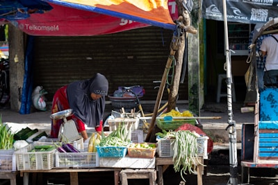 A person wearing a headscarf is tending to a variety of vegetables displayed on a wooden cart in a street market. The produce includes eggplants, green onions, and long beans, all organized in white plastic crates. The market stall is shaded by a makeshift orange and blue tarpaulin. A closed shop with a shutter is in the background.