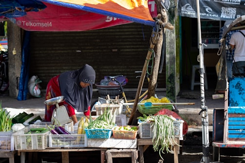 A person wearing a headscarf is tending to a variety of vegetables displayed on a wooden cart in a street market. The produce includes eggplants, green onions, and long beans, all organized in white plastic crates. The market stall is shaded by a makeshift orange and blue tarpaulin. A closed shop with a shutter is in the background.
