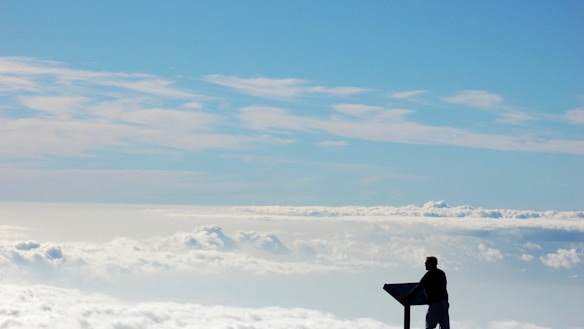 A person is standing by a viewing platform or sign overlooking a vast sky filled with light, fluffy clouds. The horizon shows layers of cloud formations under a clear blue sky, creating a serene and expansive view.