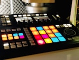 A music production controller with an array of brightly lit, colorful pads arranged in a grid. These pads are surrounded by various buttons and knobs for controlling music software. The background is slightly blurred, highlighting the focus on the controller.