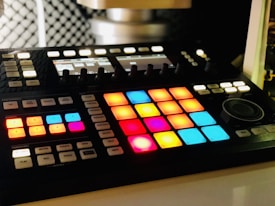 A music production controller with an array of brightly lit, colorful pads arranged in a grid. These pads are surrounded by various buttons and knobs for controlling music software. The background is slightly blurred, highlighting the focus on the controller.