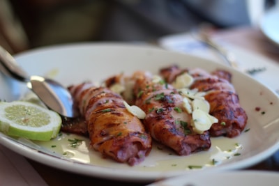 Three pieces of grilled and garnished seafood are arranged on a white plate, accompanied by a few slices of lime on the side. The dish is topped with chopped herbs and thinly sliced garlic, with a spoon positioned next to the food.