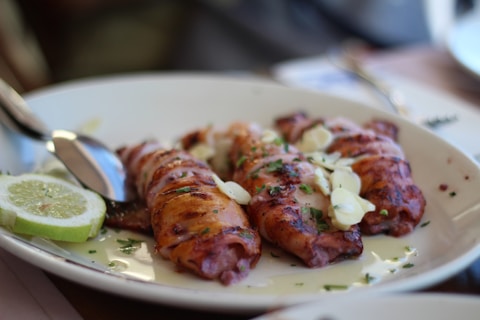 Three pieces of grilled and garnished seafood are arranged on a white plate, accompanied by a few slices of lime on the side. The dish is topped with chopped herbs and thinly sliced garlic, with a spoon positioned next to the food.