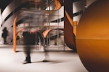 A modern interior setting featuring an abstract architectural design with curved and angular elements. The structure incorporates a mix of wooden textures and smooth surfaces, and there is a sense of motion blurred human figures passing through.