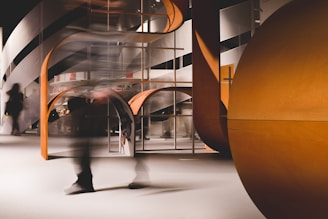 A modern interior setting featuring an abstract architectural design with curved and angular elements. The structure incorporates a mix of wooden textures and smooth surfaces, and there is a sense of motion blurred human figures passing through.