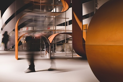 A modern interior setting featuring an abstract architectural design with curved and angular elements. The structure incorporates a mix of wooden textures and smooth surfaces, and there is a sense of motion blurred human figures passing through.