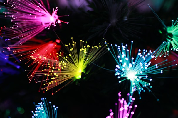 Vibrant and colorful fibers of light radiate in various directions, creating an intricate display. The fibers are primarily pink, red, yellow, and blue, their tips glowing brightly against a dark background, producing a mesmerizing and abstract visual effect.