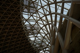 A geometric ceiling structure with crisscrossing metal beams forms a lattice pattern. The structure is predominantly glass, allowing natural light to filter through, creating an airy and modern atmosphere. The design suggests architectural sophistication and innovation.