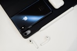 On a clean surface, there is a sleek black smartphone with a reflective Apple logo, a pair of white wireless earbuds, a matching white charging case, and a stylus pen. The setup is organized and minimalist, creating a tech-focused aesthetic.