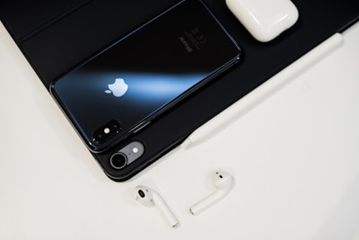 On a clean surface, there is a sleek black smartphone with a reflective Apple logo, a pair of white wireless earbuds, a matching white charging case, and a stylus pen. The setup is organized and minimalist, creating a tech-focused aesthetic.