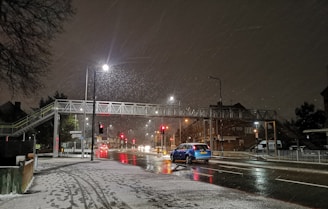 A nighttime urban scene with snow falling lightly onto a road. A pedestrian bridge spans across the street, and streetlights illuminate the wet, reflective road surface. A blue car is driving along the road, and several other vehicles and streetlights are visible in the distance. Snow has accumulated on the sidewalks and the roofs of nearby buildings.