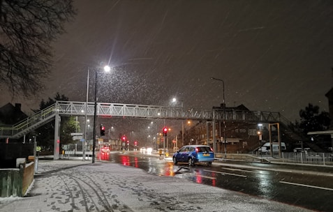 A nighttime urban scene with snow falling lightly onto a road. A pedestrian bridge spans across the street, and streetlights illuminate the wet, reflective road surface. A blue car is driving along the road, and several other vehicles and streetlights are visible in the distance. Snow has accumulated on the sidewalks and the roofs of nearby buildings.