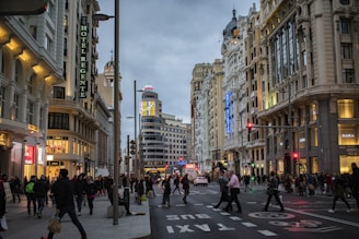 A bustling urban street scene featuring ornate, historic buildings with illuminated signage. Crowds of people walk along the sidewalks and cross the roads, while shops display their brightly lit storefronts. The sky is overcast, adding a muted backdrop to the lively city atmosphere.
