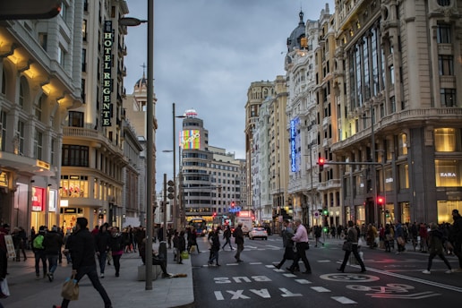 A bustling urban street scene featuring ornate, historic buildings with illuminated signage. Crowds of people walk along the sidewalks and cross the roads, while shops display their brightly lit storefronts. The sky is overcast, adding a muted backdrop to the lively city atmosphere.