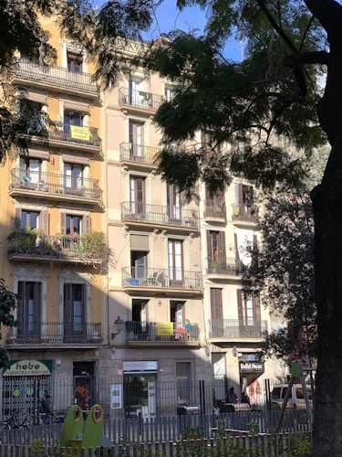 A multi-story residential building with ornate balconies and shuttered windows, partially shaded by trees. The ground floor has small commercial establishments, including a cosmetics store and a business with signage. The building is painted in light pastel colors, and balconies feature potted plants.