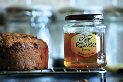 A jar of Rowse honey sits on a wire rack next to a loaf of brown bread. The honey jar is clear with a black lid, showcasing its amber contents. The bread appears freshly baked, with a textured crust and hints of raisins. The background is slightly blurred, featuring glass containers.
