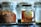 A jar of Rowse honey sits on a wire rack next to a loaf of brown bread. The honey jar is clear with a black lid, showcasing its amber contents. The bread appears freshly baked, with a textured crust and hints of raisins. The background is slightly blurred, featuring glass containers.