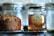 A jar of Rowse honey sits on a wire rack next to a loaf of brown bread. The honey jar is clear with a black lid, showcasing its amber contents. The bread appears freshly baked, with a textured crust and hints of raisins. The background is slightly blurred, featuring glass containers.