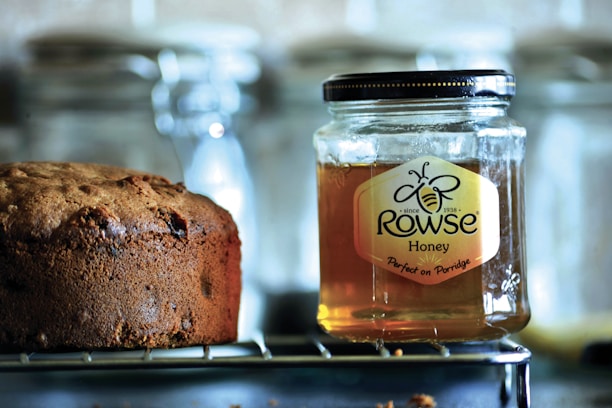 A jar of Rowse honey sits on a wire rack next to a loaf of brown bread. The honey jar is clear with a black lid, showcasing its amber contents. The bread appears freshly baked, with a textured crust and hints of raisins. The background is slightly blurred, featuring glass containers.