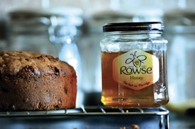 A jar of Rowse honey sits on a wire rack next to a loaf of brown bread. The honey jar is clear with a black lid, showcasing its amber contents. The bread appears freshly baked, with a textured crust and hints of raisins. The background is slightly blurred, featuring glass containers.