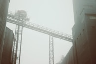 A misty industrial scene featuring large buildings and a metal framework structure, creating a sense of scale and infrastructure. The haziness adds an atmospheric and somewhat mysterious quality.