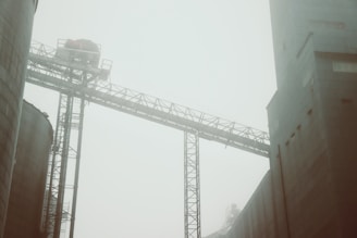 A misty industrial scene featuring large buildings and a metal framework structure, creating a sense of scale and infrastructure. The haziness adds an atmospheric and somewhat mysterious quality.