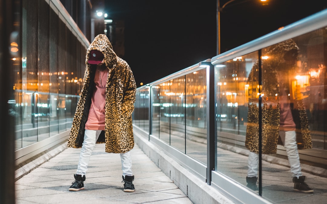 man wearing pink shirt, white pants, black high-top sneakers and brown leopard print hooded coat at night