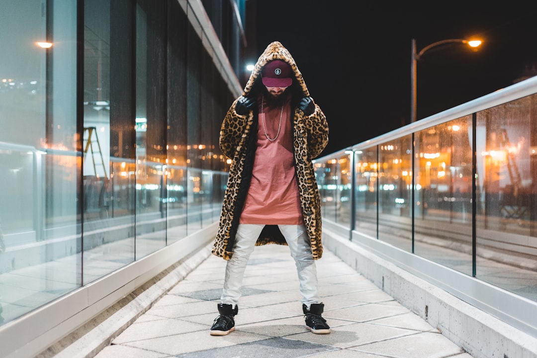 man wearing brown leopard print hooded coat, pink shirt, whit epants and black high-top sneakers beside glass building at night
