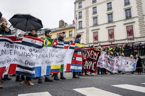 A group of people is participating in a street protest, holding banners with various flags and messages in different languages. The crowd is dressed in winter clothing, and some individuals are holding umbrellas. The background features a building with balconies and posters, and there is a gathering of onlookers and security personnel on the sidewalk.