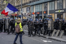 A protestor wearing a yellow vest and a helmet stands facing a line of police officers equipped with riot gear and shields. Several officers are in a defensive stance, and the protestor holds a French flag. Behind the scene, there are storefronts and a bus passing by.