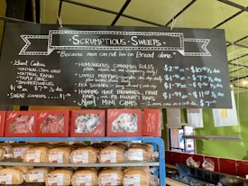 A bakery display featuring a blackboard menu with handwritten text outlining various baked goods and their prices. Below the menu are shelves stocked with bread and packaged pastries. The menu includes items like giant cookies, muffins, scones, cinnamon rolls, and brownies. The setting has a colorful and inviting atmosphere with green walls and a well-lit interior.