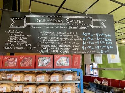A bakery display featuring a blackboard menu with handwritten text outlining various baked goods and their prices. Below the menu are shelves stocked with bread and packaged pastries. The menu includes items like giant cookies, muffins, scones, cinnamon rolls, and brownies. The setting has a colorful and inviting atmosphere with green walls and a well-lit interior.