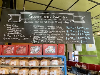 A bakery display featuring a blackboard menu with handwritten text outlining various baked goods and their prices. Below the menu are shelves stocked with bread and packaged pastries. The menu includes items like giant cookies, muffins, scones, cinnamon rolls, and brownies. The setting has a colorful and inviting atmosphere with green walls and a well-lit interior.