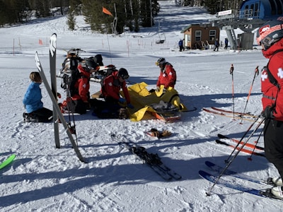 Several ski patrol members in red jackets are attending to a person on a snowy slope. There is a snowmobile nearby and various skiing equipment scattered around. The scene is set in a ski resort area with snow-covered mountains and a ski lift in the background.