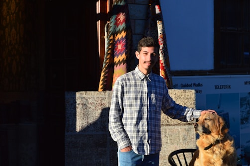 A young man stands next to a golden retriever in front of a rustic, colorful backdrop featuring traditional woven patterns. The man wears a checked shirt and jeans, gently resting his hand on the dog's head. Both are positioned in such a way that they are bathed in warm sunlight, creating a stark contrast with the shaded background. The scene includes elements of an outdoor setting like masonry and a partial view of a sign with inscriptions.